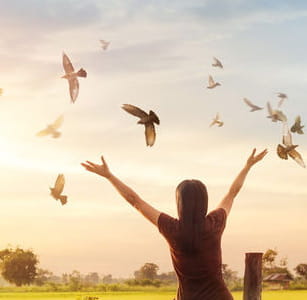 Una donna in un prato al tramonto, di spalle e con le braccia aperte verso il cielo, dove vola uno stormo di uccelli
